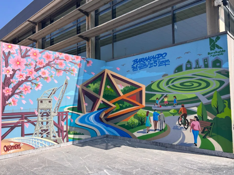 Mural en esquina con un cerezo en flor, una grúa y una pasarela, un río que sale de un poliedro y un parque con gente paseando y en bici