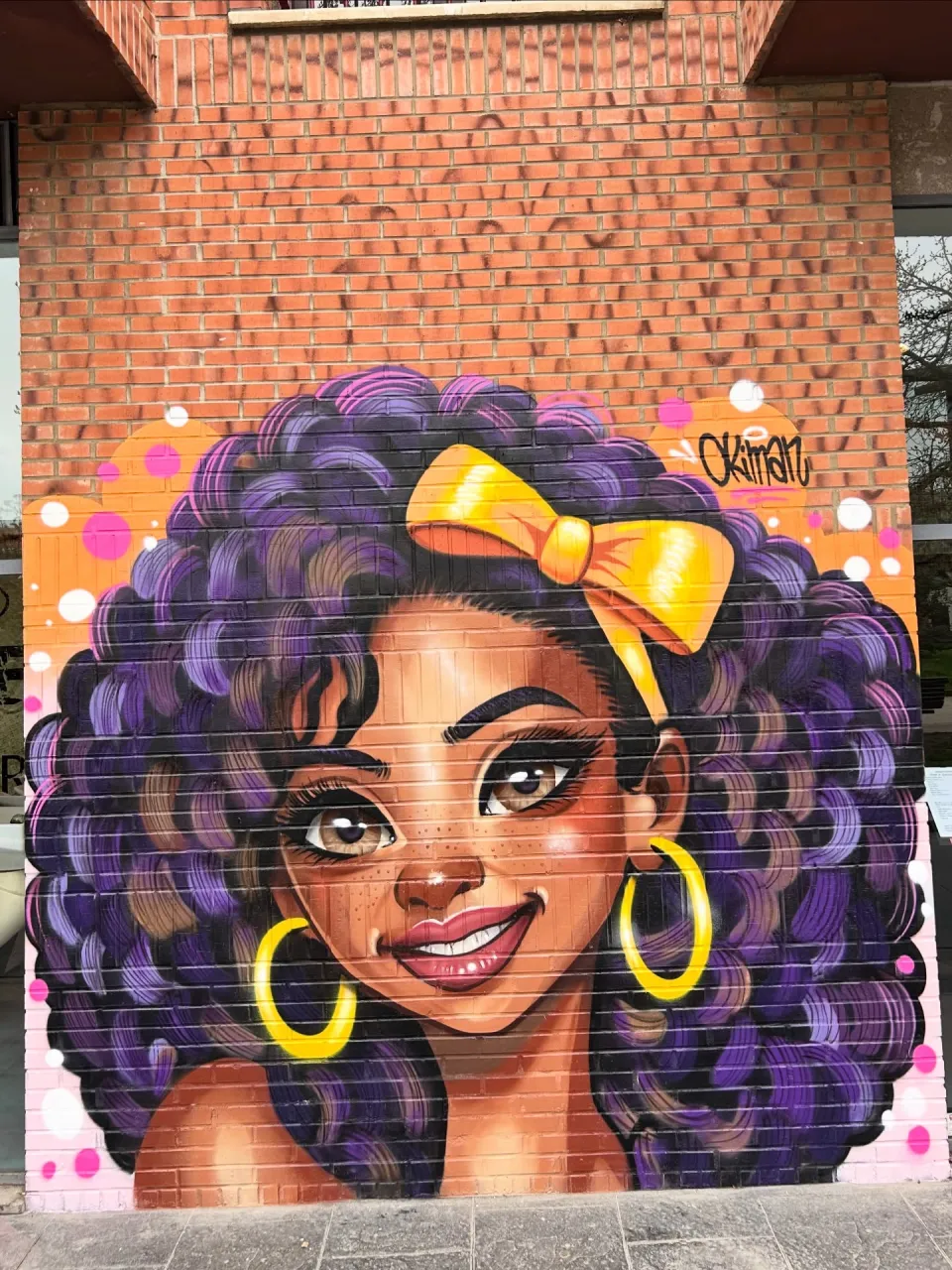 Persiana pintada con el retrato de una mujer de pelo rizado con un lazo amarillo, junto a un muro de ladrillo